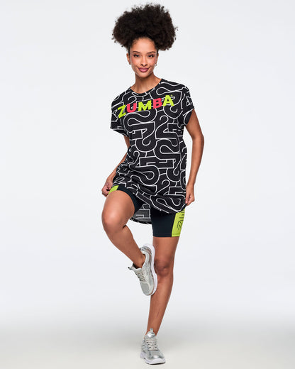 Black, long, round hem tee with an allover white 25 graphic and multicolor Zumba logo across the chest. A female model wears the round hem tee, black bike shorts with neon green side panels and silver sneakers against a light gray background.