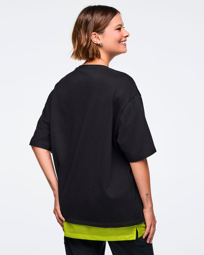 Black oversized Zumba T-shirt shown from the back, with an extended double layered mesh hem panel. Female model is turned sideways and wearing the loose short-sleeve tee with black bottoms.