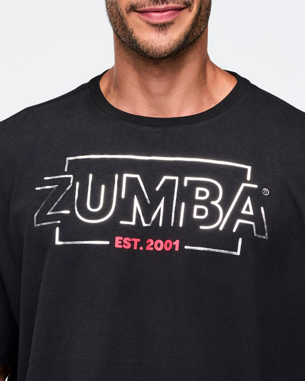 Black oversized Zumba T-shirt with a large silver ZUMBA logo and pink “EST. 2001” text on the front. Male model is wearing the loose short-sleeve tee in a close-up view highlighting the chest graphic and crew neckline.