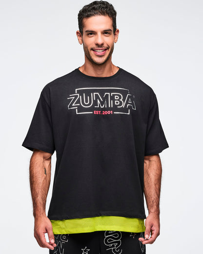 Black oversized Zumba T-shirt with a large silver ZUMBA logo and pink “EST. 2001” text on the front, with an extended double layered mesh hem panel. Male model is wearing the loose short-sleeve tee with black graphic pants.