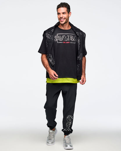 Black oversized Zumba T-shirt with a large silver ZUMBA logo and pink “EST. 2001” text on the front, with an extended double layered mesh hem panel. Male model is wearing the loose short-sleeve tee with black jogger pants, silver sneakers, and a black printed sweatshirt draped over his shoulders.
