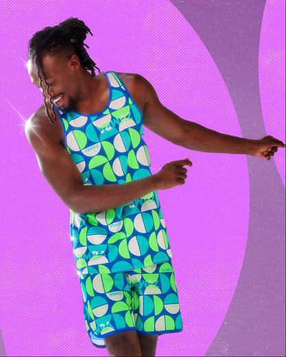 Male model in a dark blue racerback muscle tank with a geometric print of half circles in aqua, neon green, and white with 'ZUMBA' text, paired with matching print shorts, against a bright pink background with purple arcs.