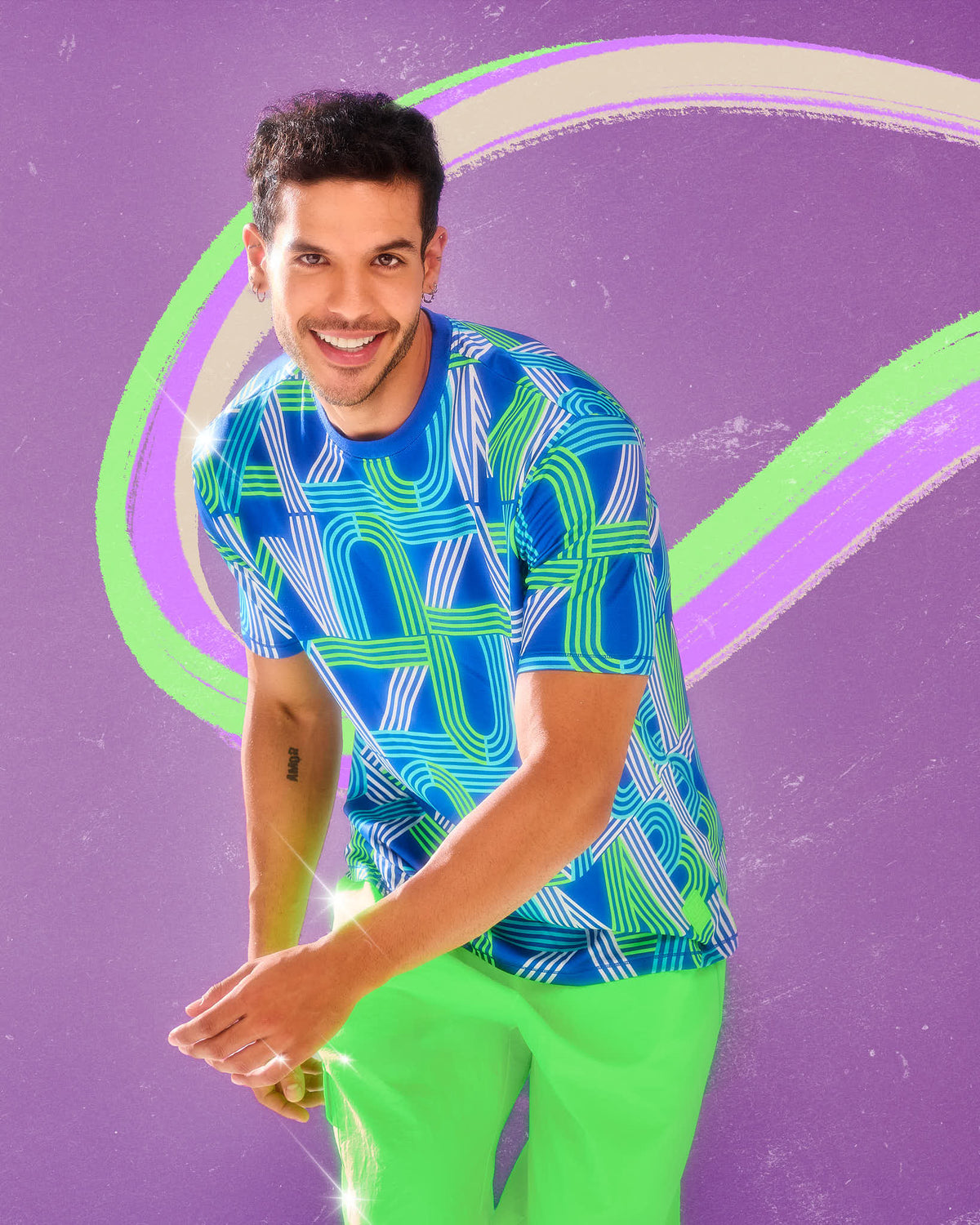Male model in a dark blue mesh top with a retro print of large 'ZUMBA' letters drawn in white, aqua, and neon green stripes, paired with neon green shorts, against a purple background with swirling green and cream stripes.