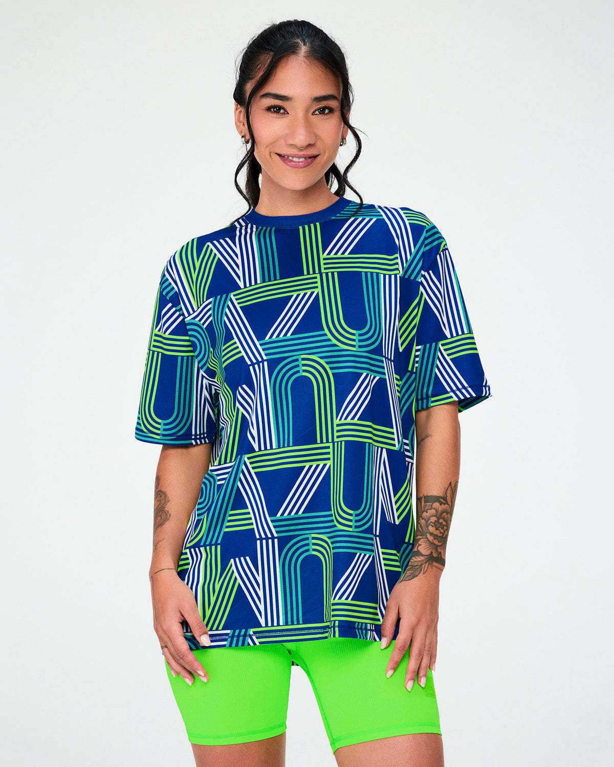 Female model in a dark blue mesh top with a retro print of large 'ZUMBA' letters drawn in white, aqua, and neon green stripes, worn loose over neon green biker shorts.