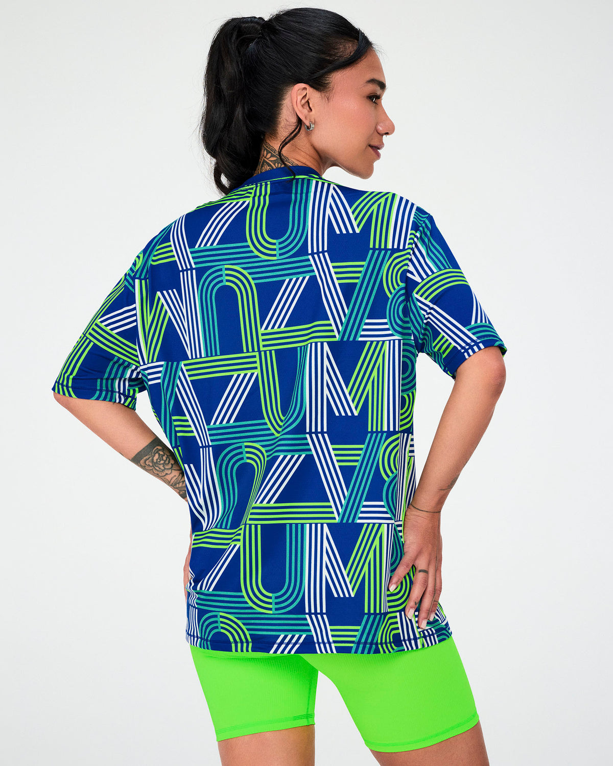 Shown from the back, a female model in the dark blue striped 'ZUMBA' letter mesh top, worn oversized with neon green biker shorts.