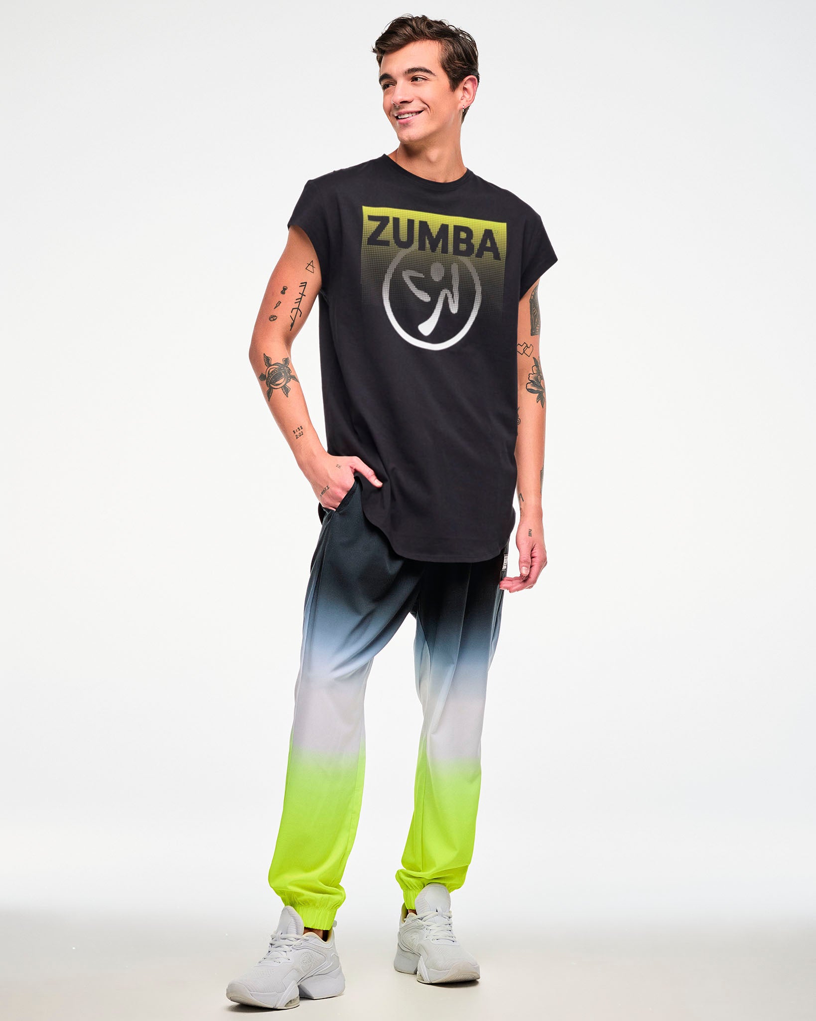 Male model wearing a black cap-sleeve tee with a yellow-to-white gradient "ZUMBA" logo and Zumbito graphic, paired with black-to-neon-yellow ombre joggers and white sneakers.