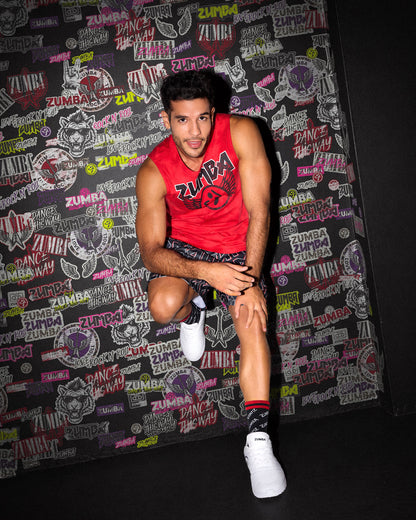 A male model crouching against a black wall covered in rock-like Zumba emblems, 
wearing a red sleeveless Zumba tank, patterned Zumba shorts and matching socks, paired with white shoes.