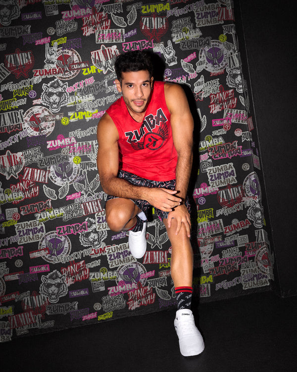 A male model crouching against a black wall covered in rock-like Zumba emblems, 
wearing a red sleeveless Zumba tank, patterned Zumba shorts and matching socks, paired with white shoes.