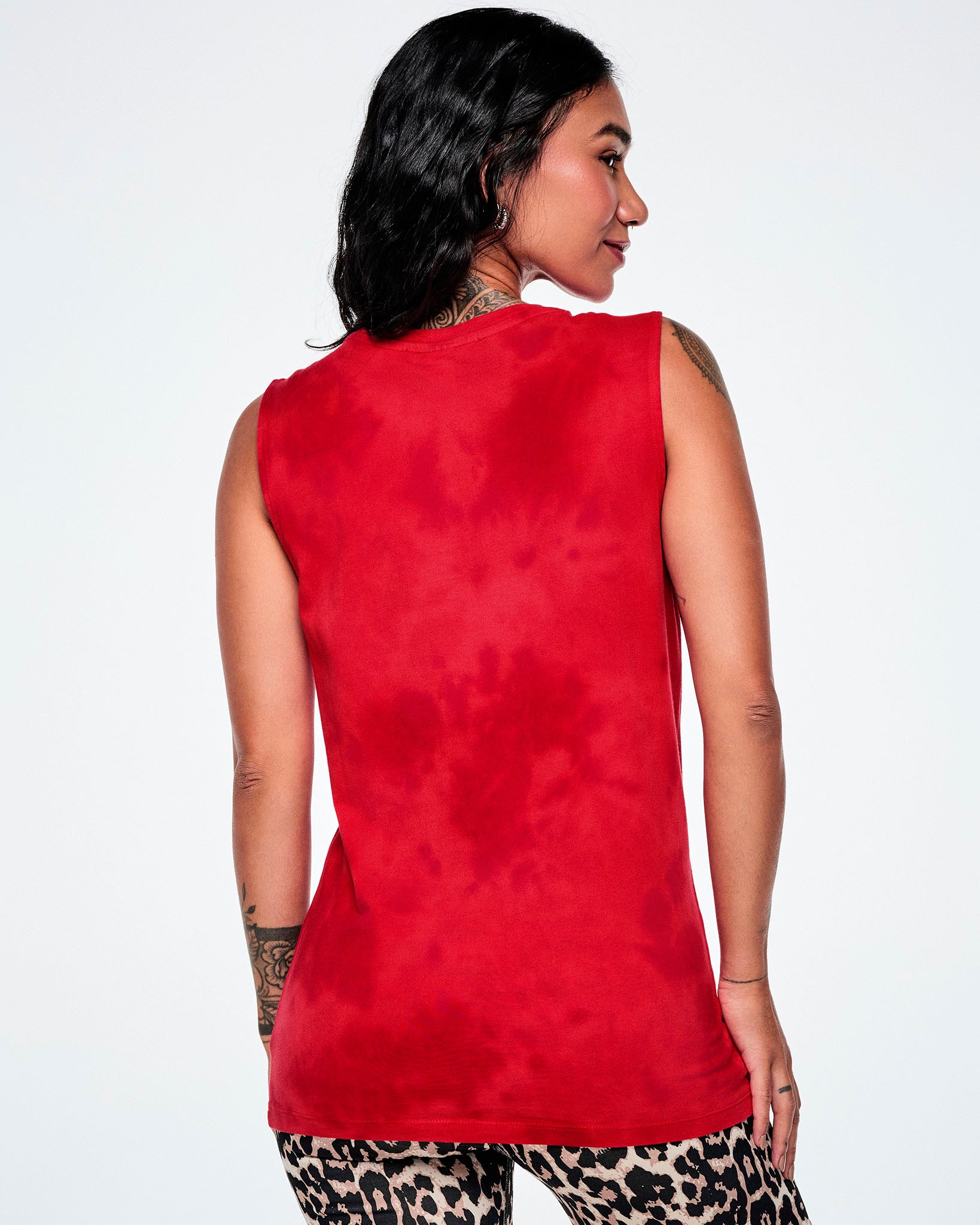 Back view of a female model wearing a red tank top with a distressed black and white Zumba text with a large black Zumbito graphic with wings, paired with tan leopard print leggings.
