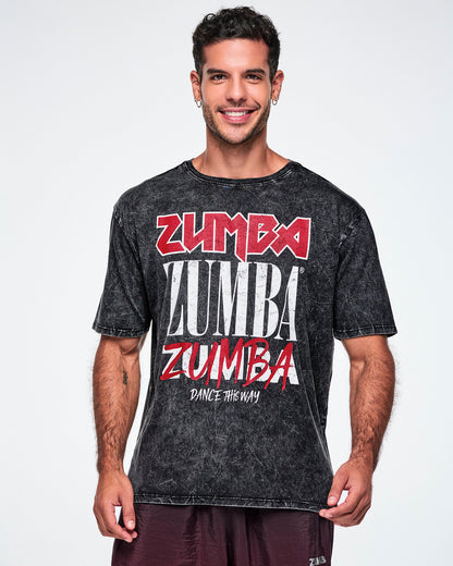 A male model wearing a black acid-wash graphic t-shirt with 'ZUMBA' printed in red and white in four different fonts, with "Dance This Way" in white text below and paired with black pants.