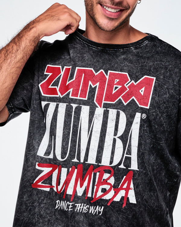 A close-up of a male model wearing a black acid-wash graphic tee featuring the word 'ZUMBA' printed in red and white multiple times, with 'DANCE THIS WAY' underneath.