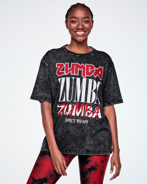 A female model wearing a black acid-wash graphic t-shirt with 'ZUMBA' printed in red and white in four different fonts, with 