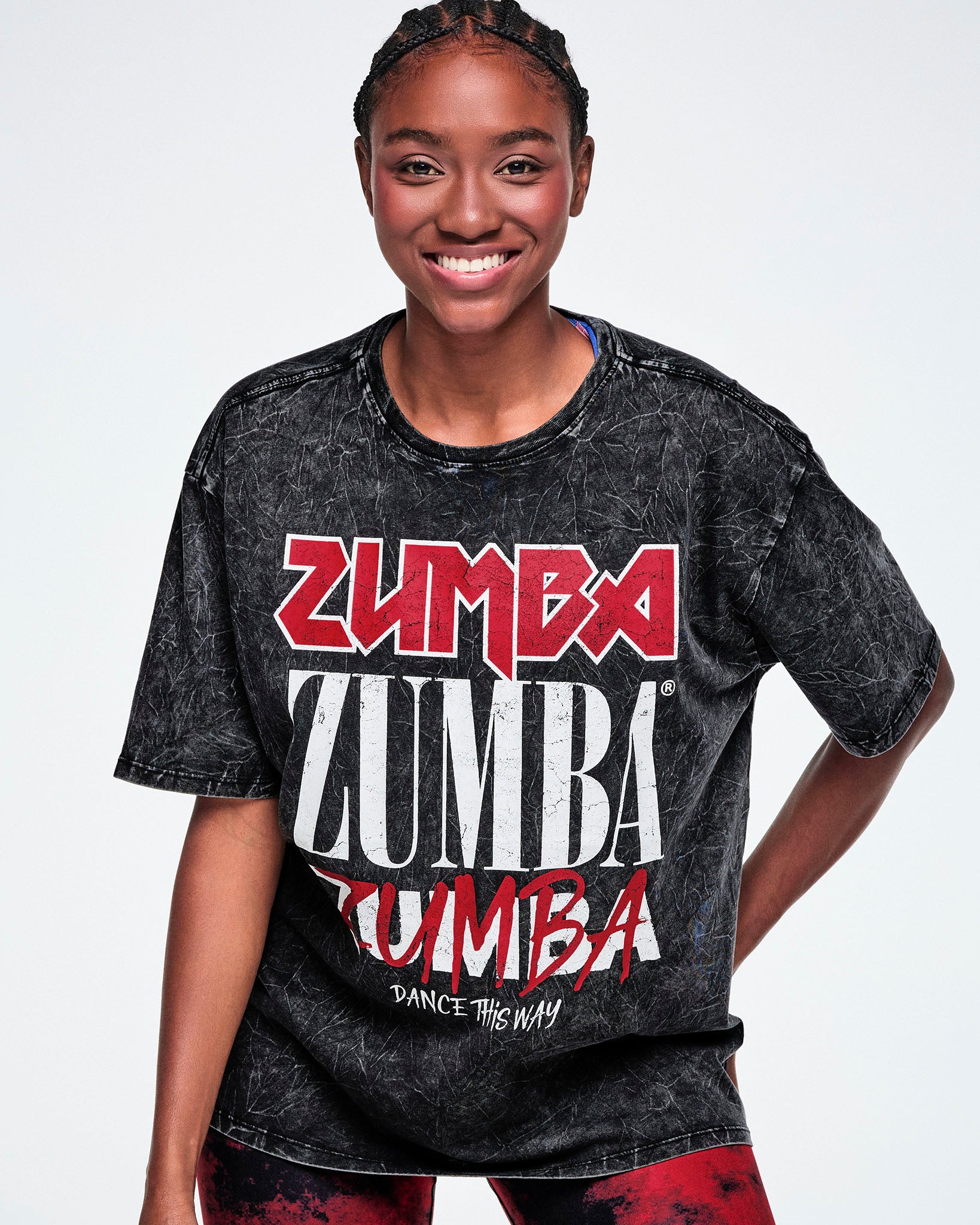 A smiling female model wearing a black acid-wash graphic tee with 'ZUMBA' printed in red and white, paired with red and black leggings, against a white background.