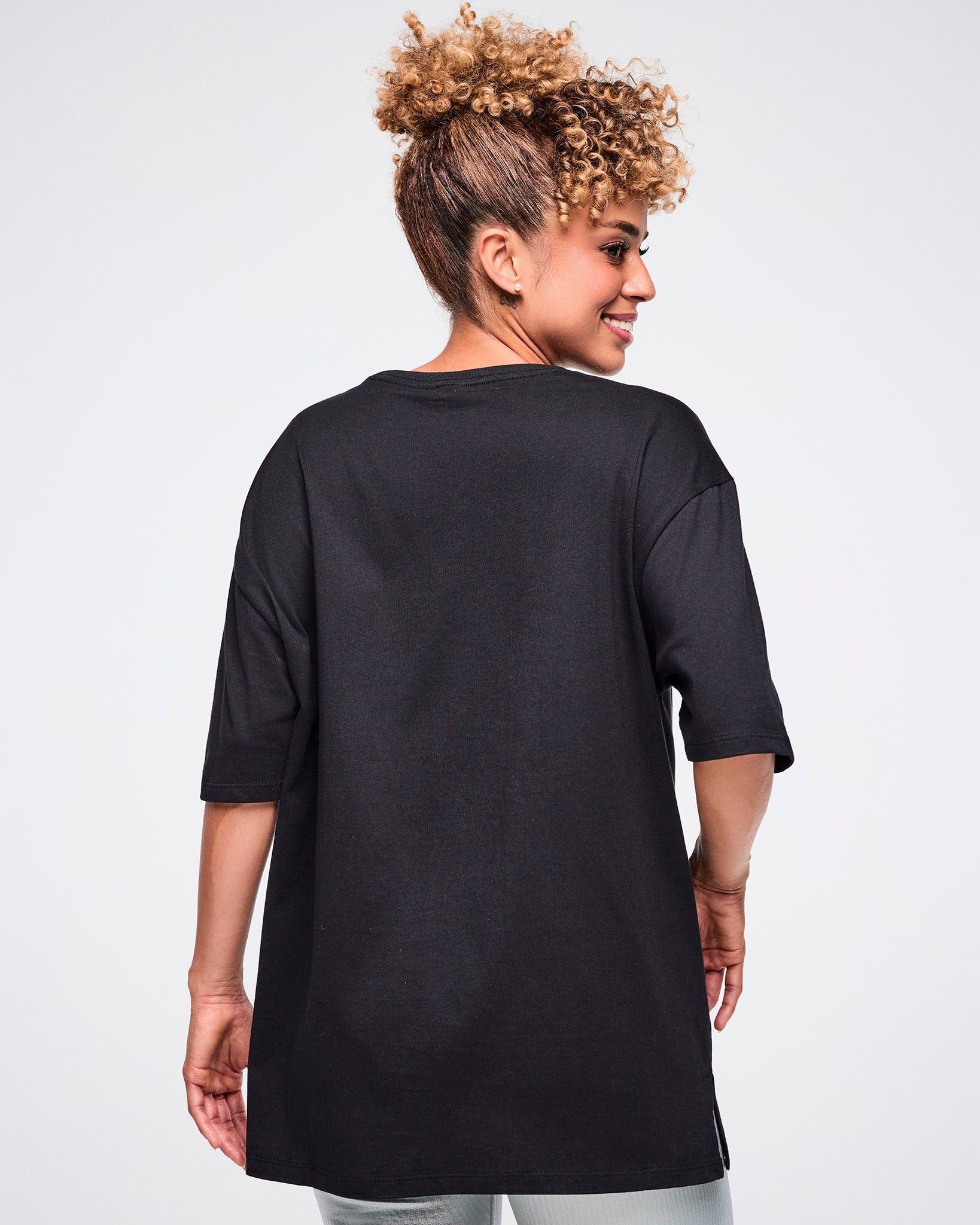 Back view of a female model wearing a black Zumba T-shirt with “ZUMBA” in a silver patterned rectangle, paired with matching pants.