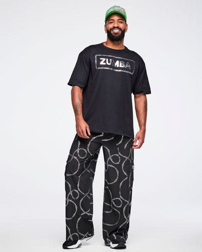 Full view of a male model wearing a black Zumba T-shirt with “ZUMBA” in a silver patterned rectangle, paired with matching pants and black and white shoes.