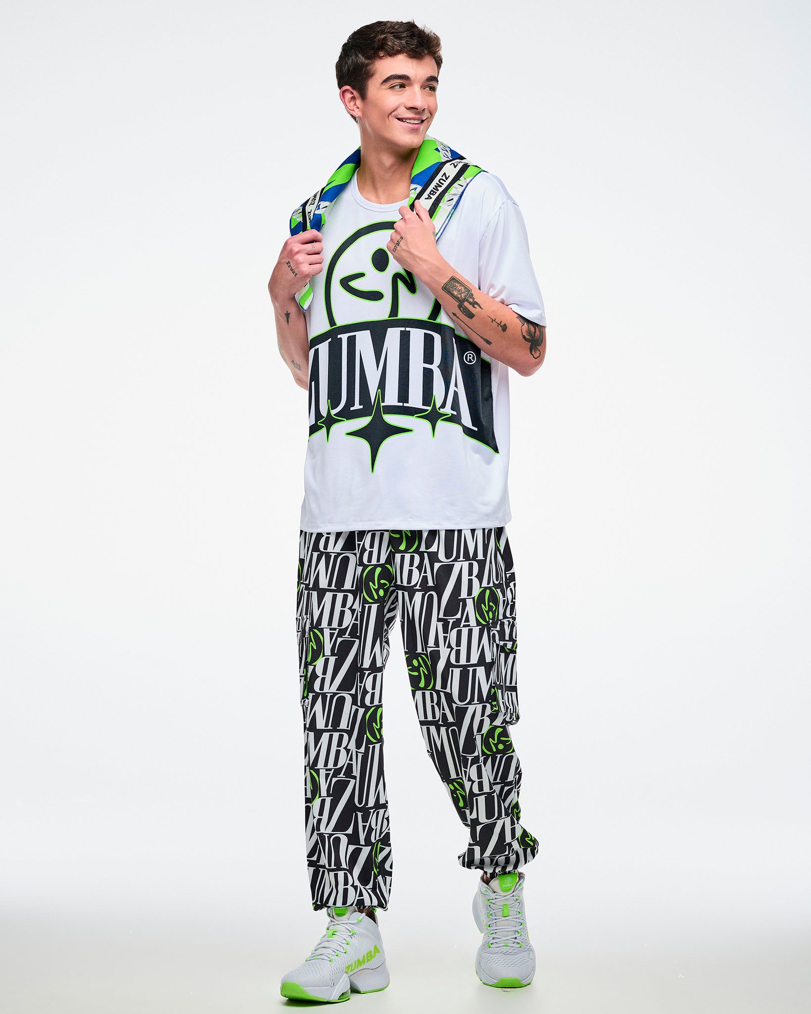 Male model holds a colorful bandana at his neck and wears a white Zumba top with a black and green logo, black‑white‑green pants, and white‑green shoes on a white background.