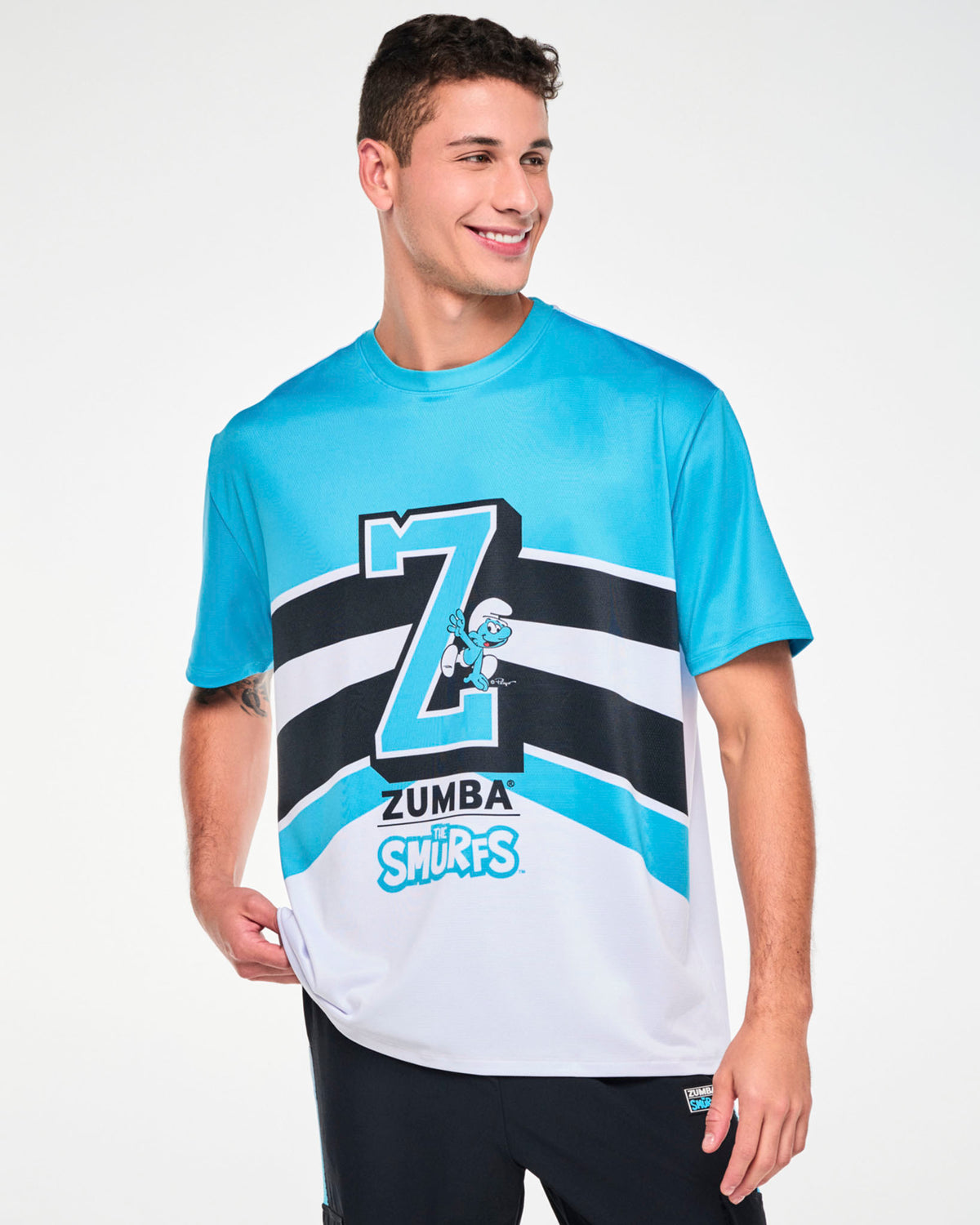 Male model in a blue and white mesh top with a large blue varsity 'Z' outlined in black and white, a Smurf climbing the letter, and 'ZUMBA' over 'THE SMURFS' logos below, all set against sweeping black and white stripes. Paired with black cargo pants.