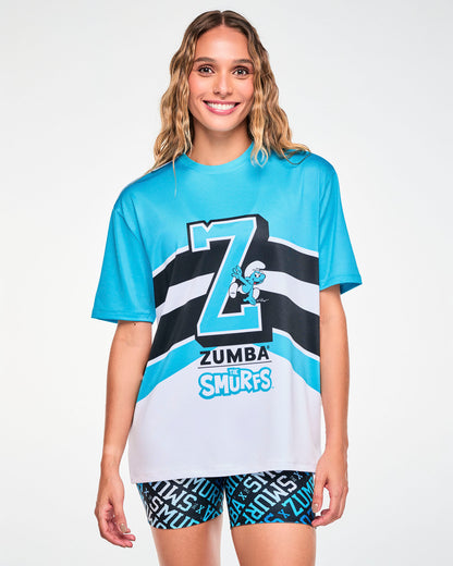 Female model in a blue and white mesh top featuring a large blue varsity 'Z' outlined in black and white with a Smurf climbing up the letter, 'ZUMBA' and 'THE SMURFS' logos beneath, and sweeping black and white stripes across the front. Paired with black and blue 'ZUMBA x THE SMURFS' print biker shorts.