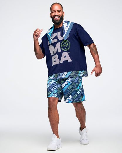Full view of a male model wearing a blue “Zumba” T-shirt, a bandana draped around his neck, paired with patterned blue “Zumba” shorts and white shoes.