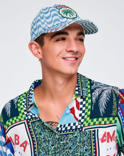 Light blue wavy-print baseball cap with embroidered “ZUMBA” text and palm-tree patch, modeled by a male wearing a multicolor printed button-up shirt on a white studio background.