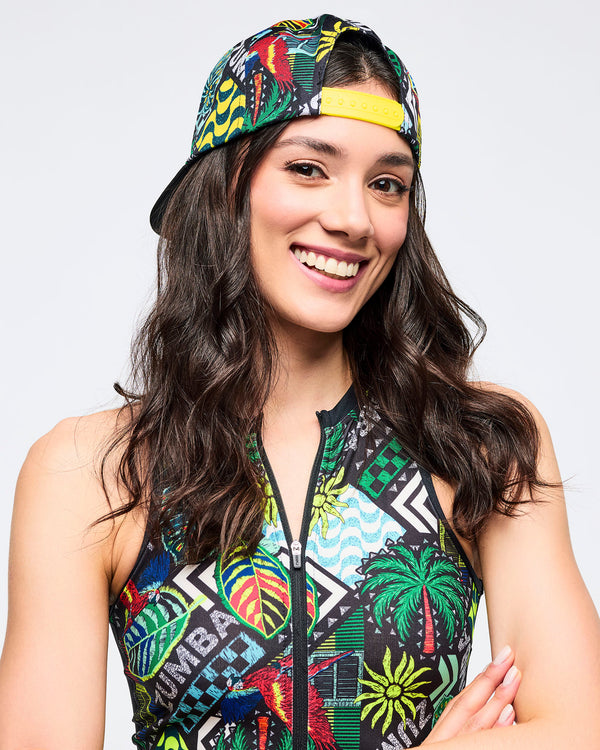 Close-up of woman wearing the cap backwards, showing the yellow snapback closure and tropical-print panels; she’s in a sleeveless zip-front bodysuit with a bold tropical/graphic print.