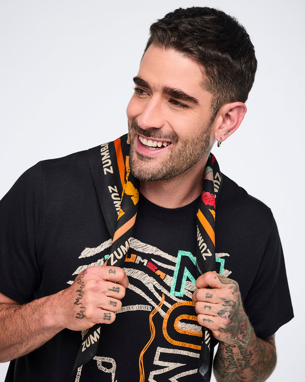 Multicolor Zumba square bandana worn loosely around the neck on a male model. He wears a black graphic Zumba T-shirt and smiles while holding the bandana ends against a light gray background.