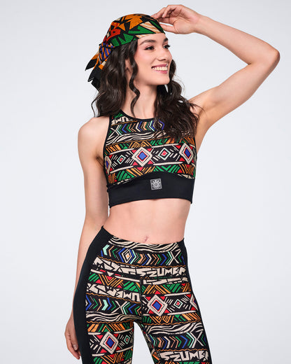 Multicolor Zumba square bandana styled as a headscarf on a female model. She wears a black printed high-neck sports bra and matching high-waisted leggings with geometric multicolor patterns.