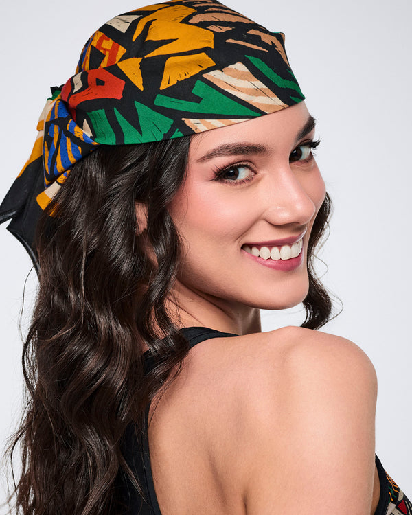 Multicolor Zumba square bandana styled as a headscarf on a female model. She wears the bandana tied over her dark wavy hair with a black sleeveless top, shown in a close-up over-the-shoulder portrait.
