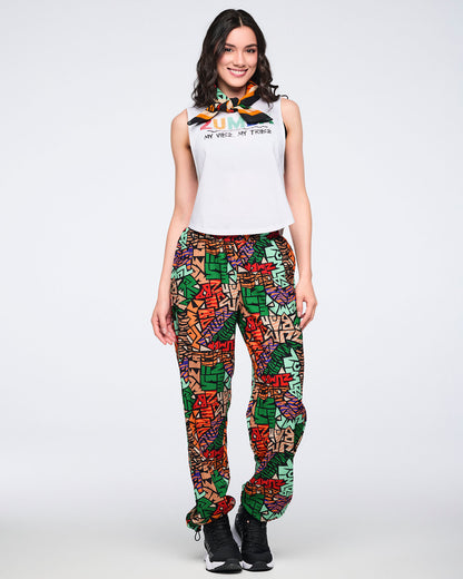 Multicolor Zumba square bandana tied at the neck on a female model. She wears a white sleeveless Zumba tank, bright abstract-print jogger pants, and black sneakers against a light gray background.