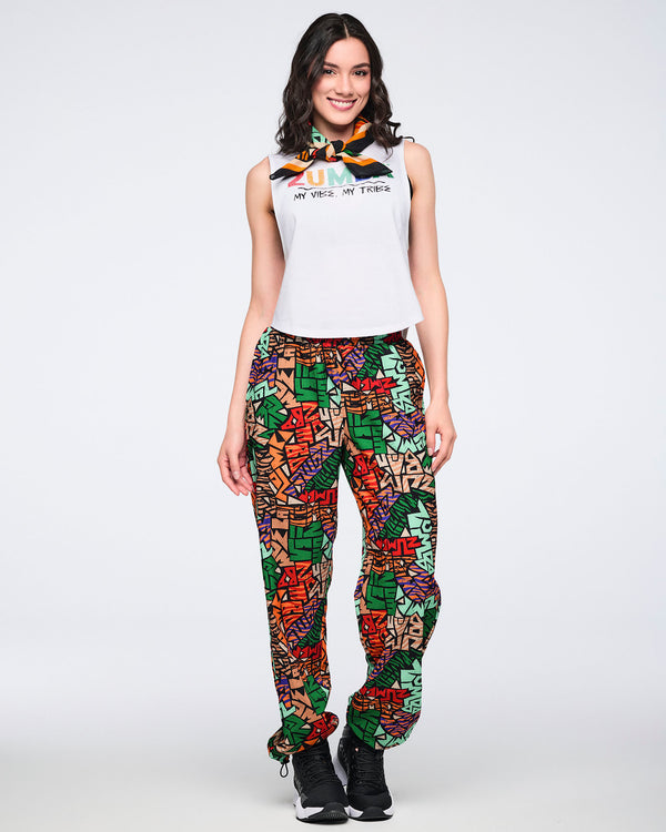 Multicolor Zumba square bandana tied at the neck on a female model. She wears a white sleeveless Zumba tank, bright abstract-print jogger pants, and black sneakers against a light gray background.