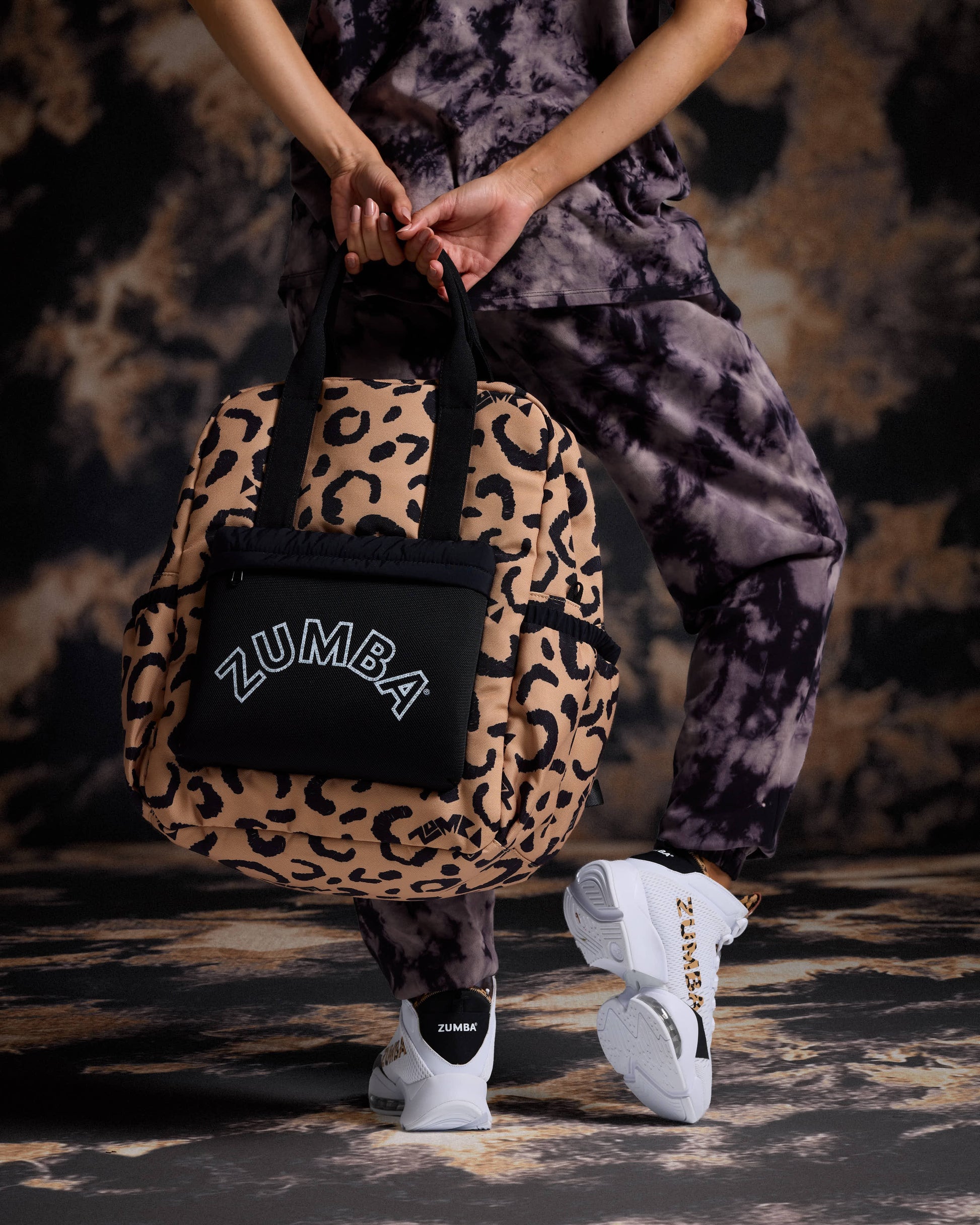 Leopard-print Zumba backpack with black top handles, side pockets, and a black front pouch featuring a white ZUMBA logo. Female model wears a black and gray tie-dye short-sleeve tee, matching tie-dye joggers, black socks, and white Zumba sneakers with leopard accents while holding the bag behind her.