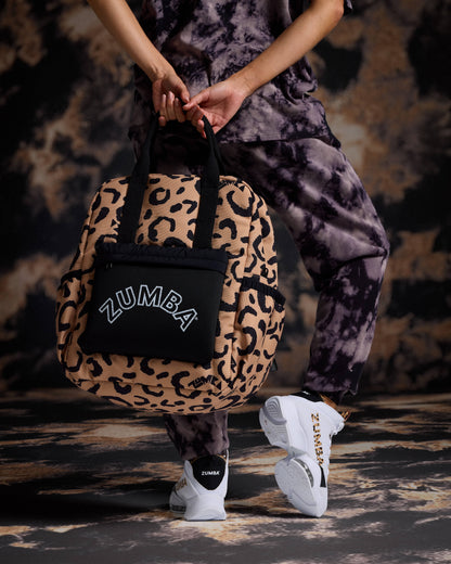 Leopard-print Zumba backpack with black top handles, side pockets, and a black front pouch featuring a white ZUMBA logo. Female model wears a black and gray tie-dye short-sleeve tee, matching tie-dye joggers, black socks, and white Zumba sneakers with leopard accents while holding the bag behind her.