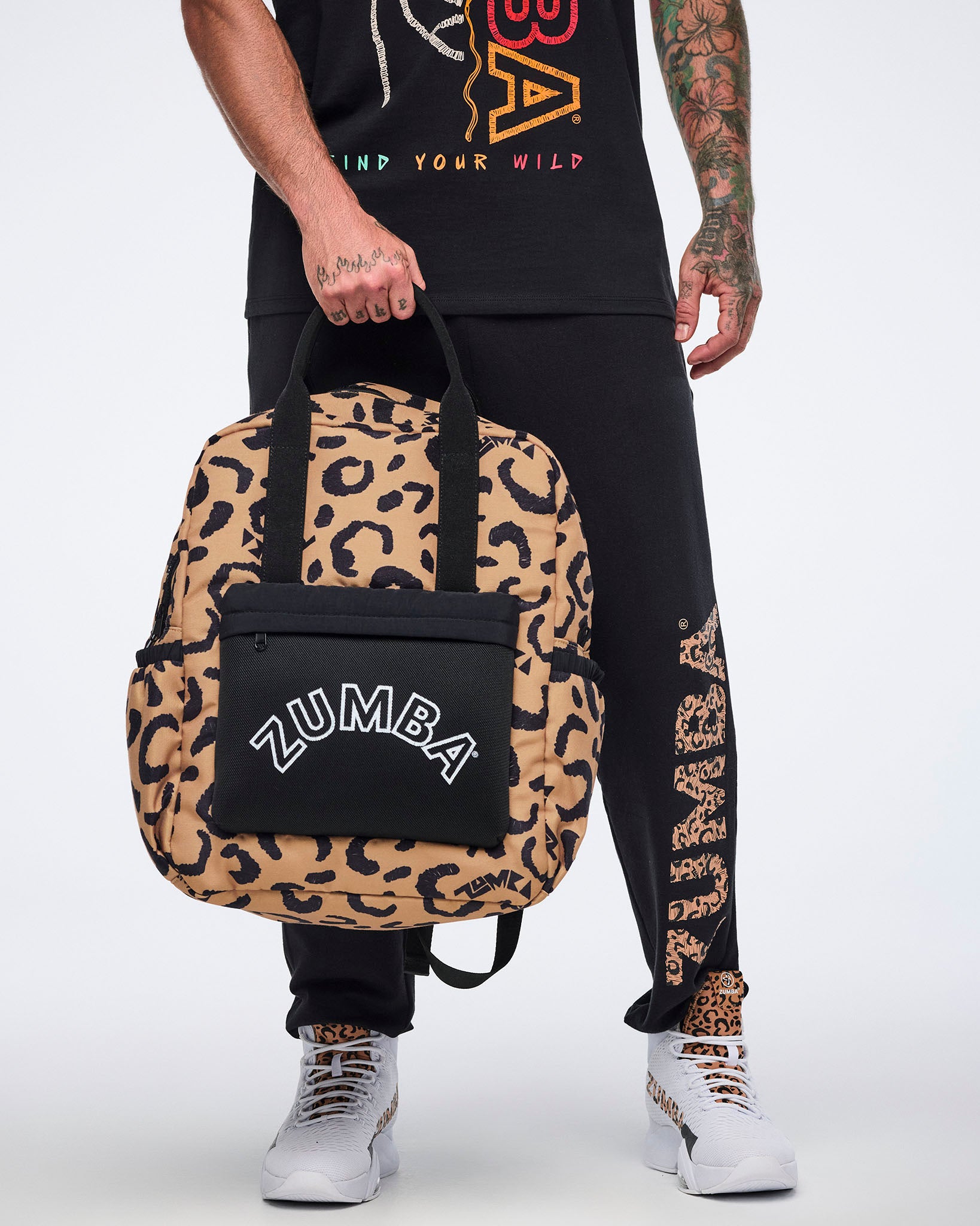 Leopard-print Zumba backpack with black handles, side pockets, and a black front pouch with white ZUMBA logo. Male model wears a black graphic Zumba T-shirt, black jogger pants with leopard-print Zumba lettering down one leg, and white high-top sneakers with leopard details while holding the bag at his side.