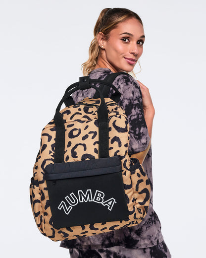 Leopard-print Zumba backpack with black handles and a black front pocket with white ZUMBA lettering, shown worn on the back. Female model wears a black and gray tie-dye short-sleeve top and matching joggers, with her hair in a ponytail, as she looks over her shoulder against a light gray background.