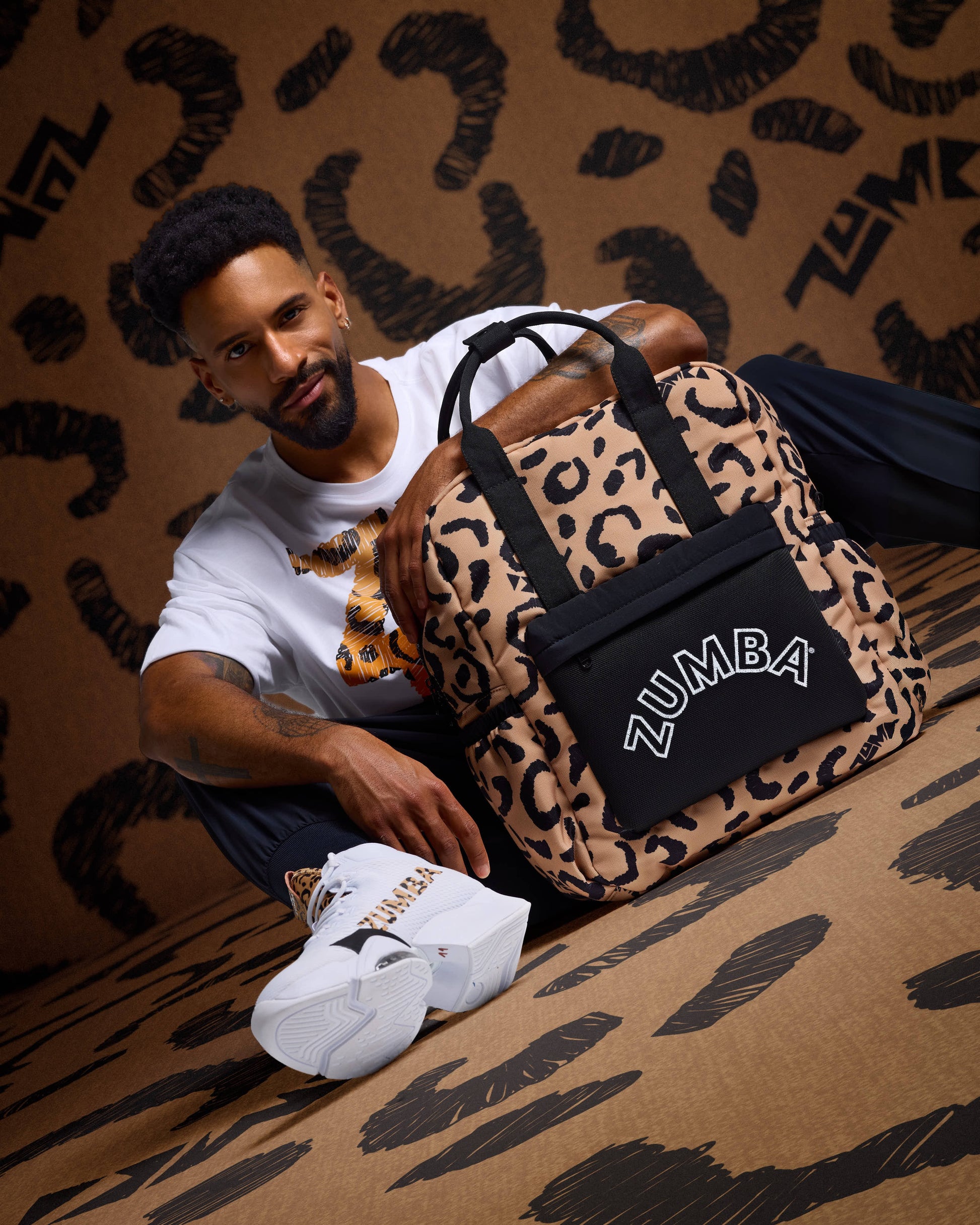 Leopard-print Zumba backpack with black top handles and a black front pouch with white ZUMBA logo, posed beside a seated male model wearing a white graphic T-shirt, black pants, and white Zumba sneakers with leopard accents against a matching leopard-print backdrop.