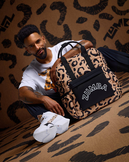 Leopard-print Zumba backpack with black top handles and a black front pouch with white ZUMBA logo, posed beside a seated male model wearing a white graphic T-shirt, black pants, and white Zumba sneakers with leopard accents against a matching leopard-print backdrop.