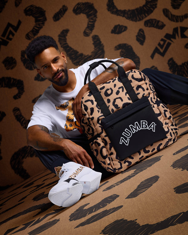 Leopard-print Zumba backpack with black top handles and a black front pouch with white ZUMBA logo, posed beside a seated male model wearing a white graphic T-shirt, black pants, and white Zumba sneakers with leopard accents against a matching leopard-print backdrop.