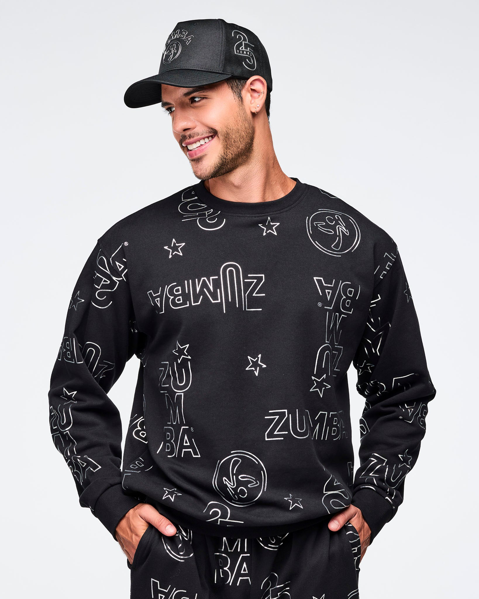 Black baseball cap with silver Zumba logo on the front and a silver 25 Years graphic on the side. A smiling male model wears the cap with a black crewneck sweatshirt covered in silver Zumba graphics and matching black joggers against a light gray background.