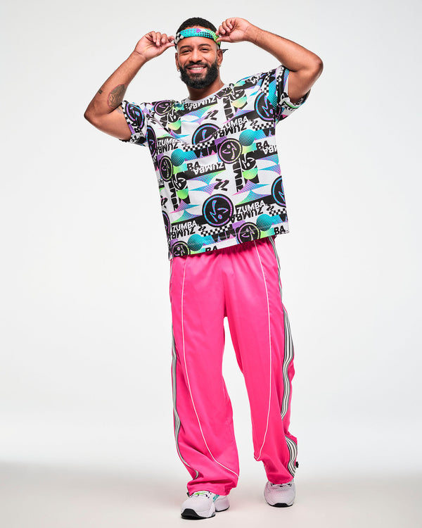 Full view of a male model wearing a bandana as a headband, a retro-print graphic tee, pink track pants with striped side panels and white shoes.
