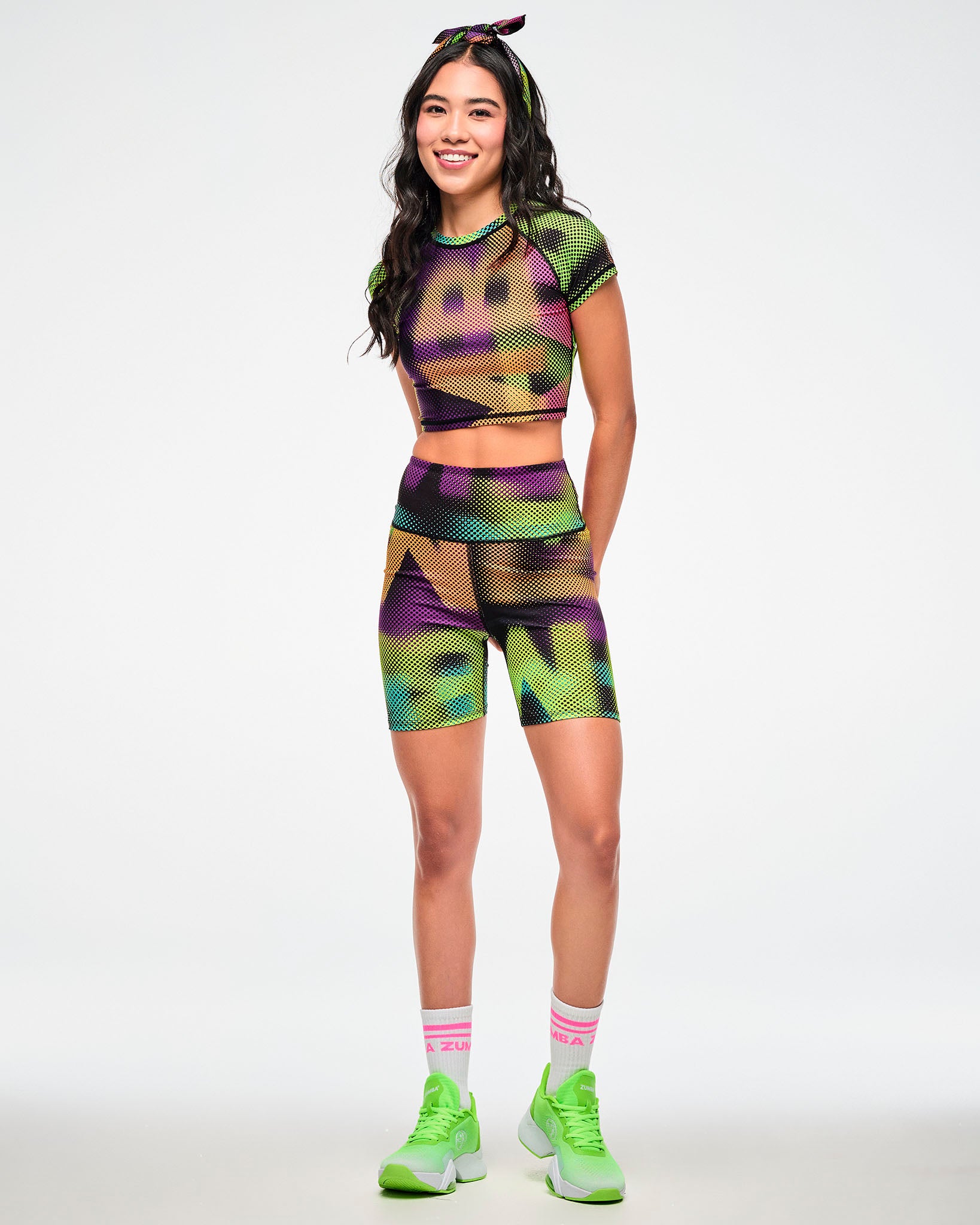 Female model wearing a colorful halftone bandana as a hair tie and paired with matching crop top, biker shorts and white shoes with white and pink "ZUMBA" socks.