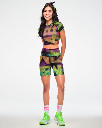 Female model wearing a colorful halftone bandana as a hair tie and paired with matching crop top, biker shorts and white shoes with white and pink "ZUMBA" socks.