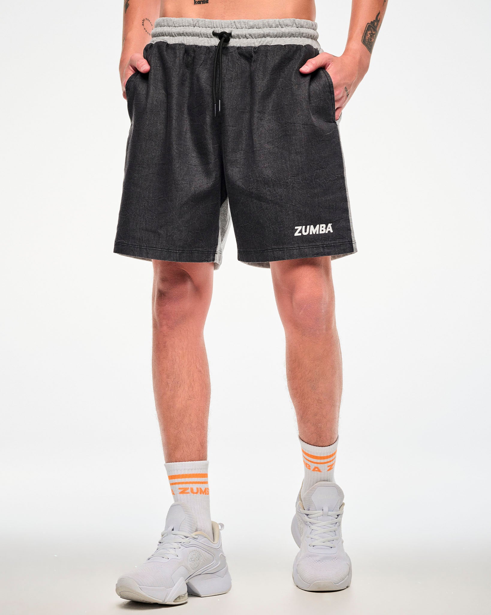 Male model wearing black and grey colorblock shorts with white "ZUMBA" text on the left leg, paired with orange-striped socks and white shoes.