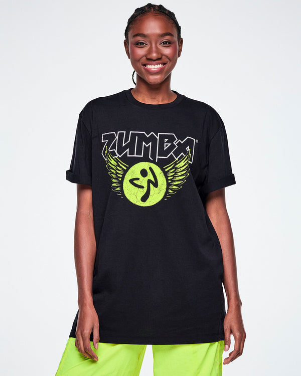 A female model modeling a black Zumba t-shirt with a large graphic on the chest featuring the word 'ZUMBA' in white outlined rock-like letters, a yellow-green circular Zumbitio with wings, paired with Zumba green pants.