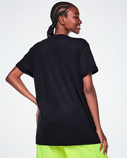 A female model shown from the back wearing a black short-sleeve tee and Zumba green pants.