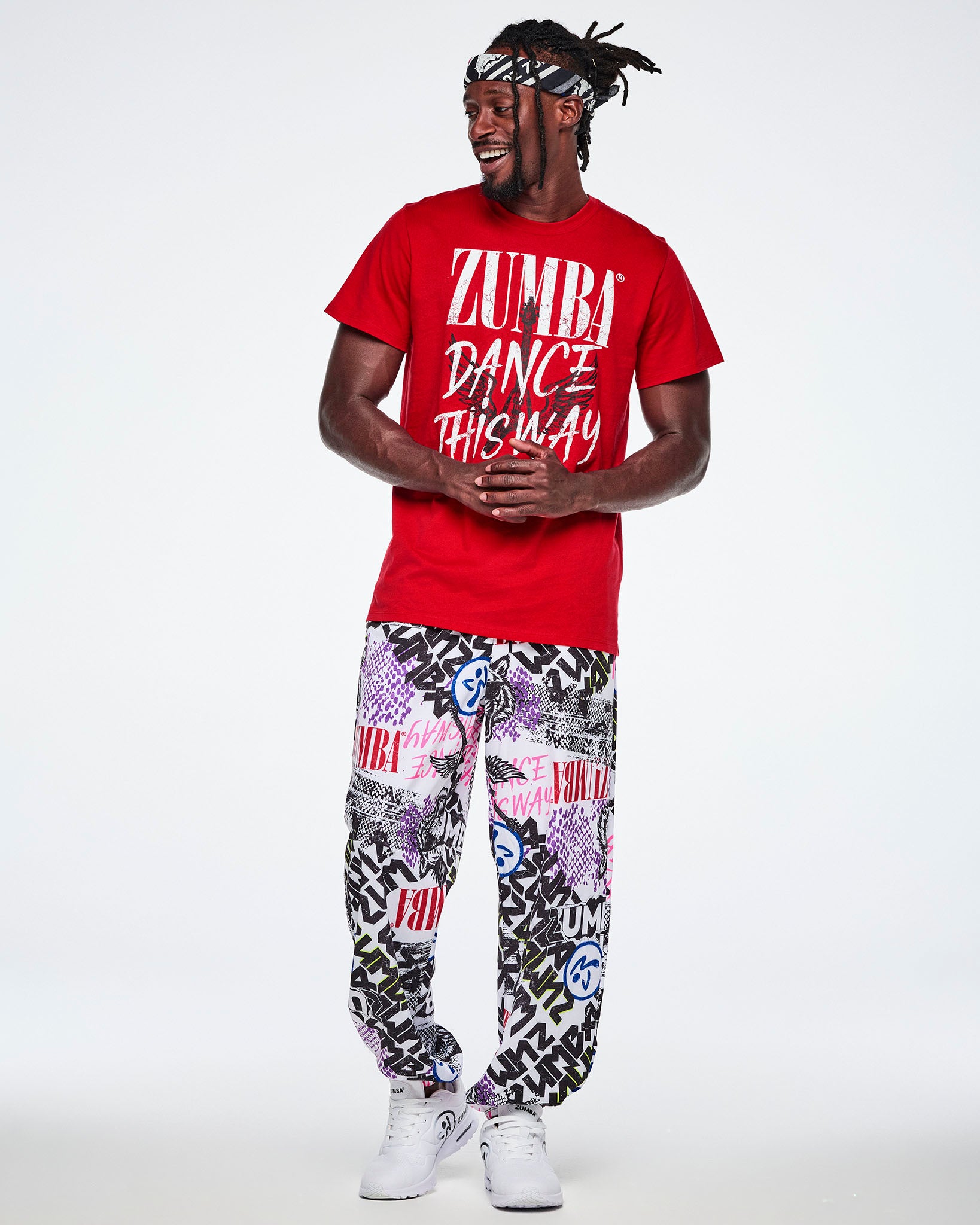 Full view of a male model wearing a bandana as a headband, a red Zumba graphics rock tee with white text 'ZUMBA DANCE LIKE THIS WAY' with a black guitar with wings in the background and paired with graphic white pants and white shoes.