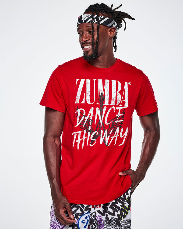 A male model wearing a bandana as a headband while wearing a red Zumba graphics rock tee that reads 'ZUMBA DANCE LIKE THIS WAY' with a black guitar with wings in the background.