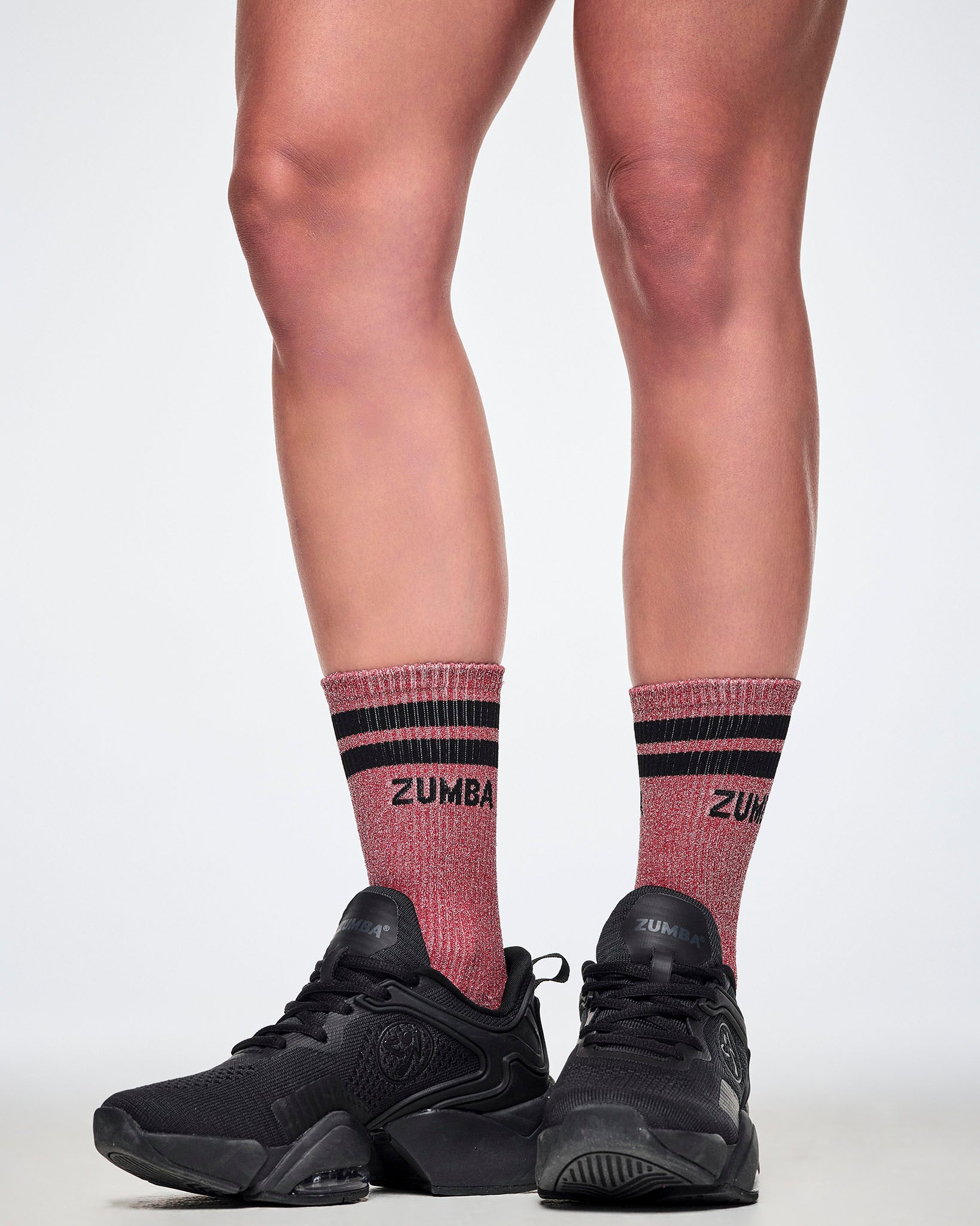 A female model wearing a pair of red high socks with black stripes, with 'ZUMBA' printed in black on the upper cuff and the sparkling lurex threads seen all-over and paired with black shoes.