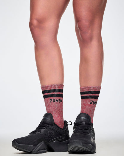 A female model wearing a pair of red high socks with black stripes, with 'ZUMBA' printed in black on the upper cuff and the sparkling lurex threads seen all-over and paired with black shoes.