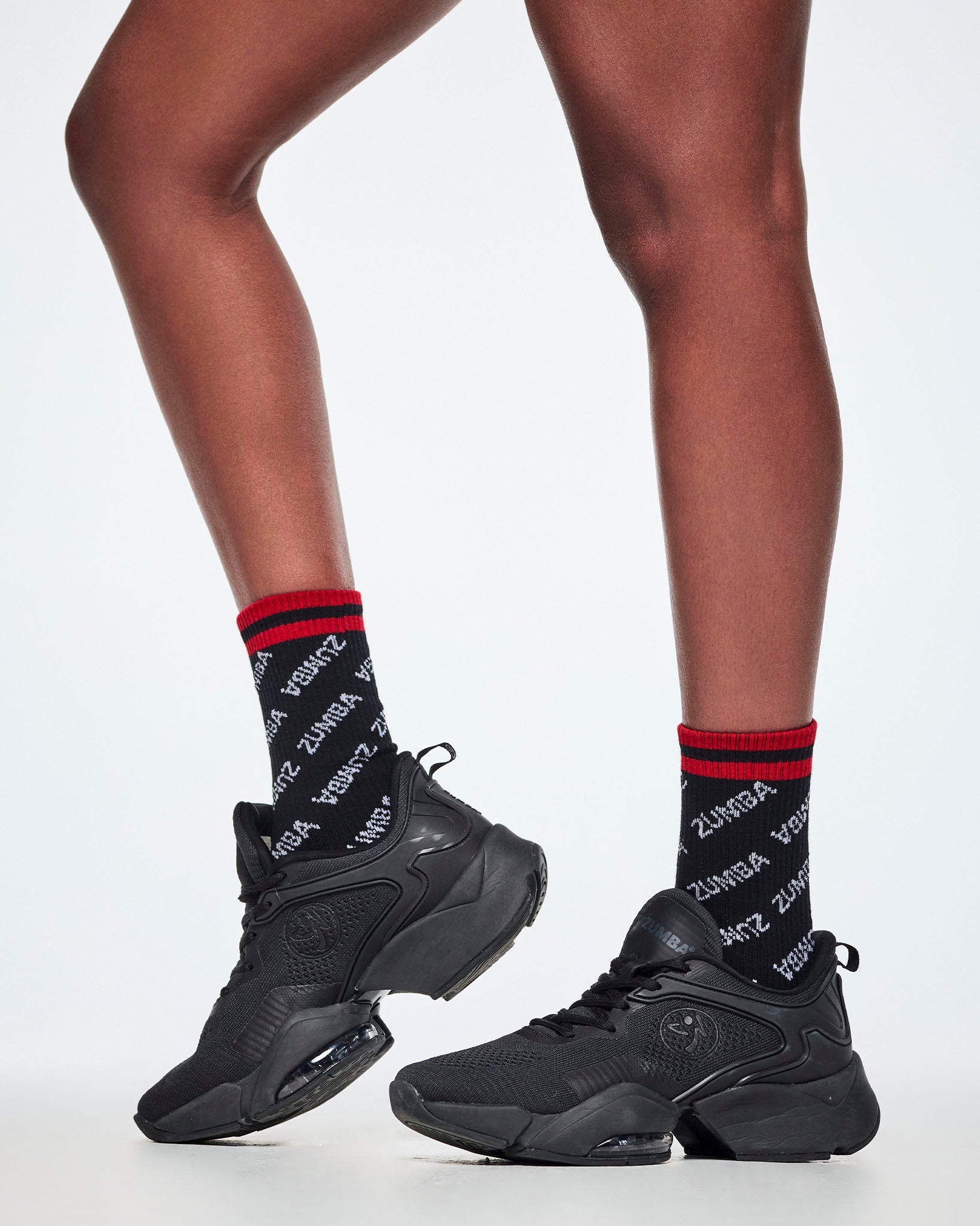 A female model wearing a pair of tube socks with a black base, white 'ZUMBA' text pattern and red cuffs, paired with black shoes.