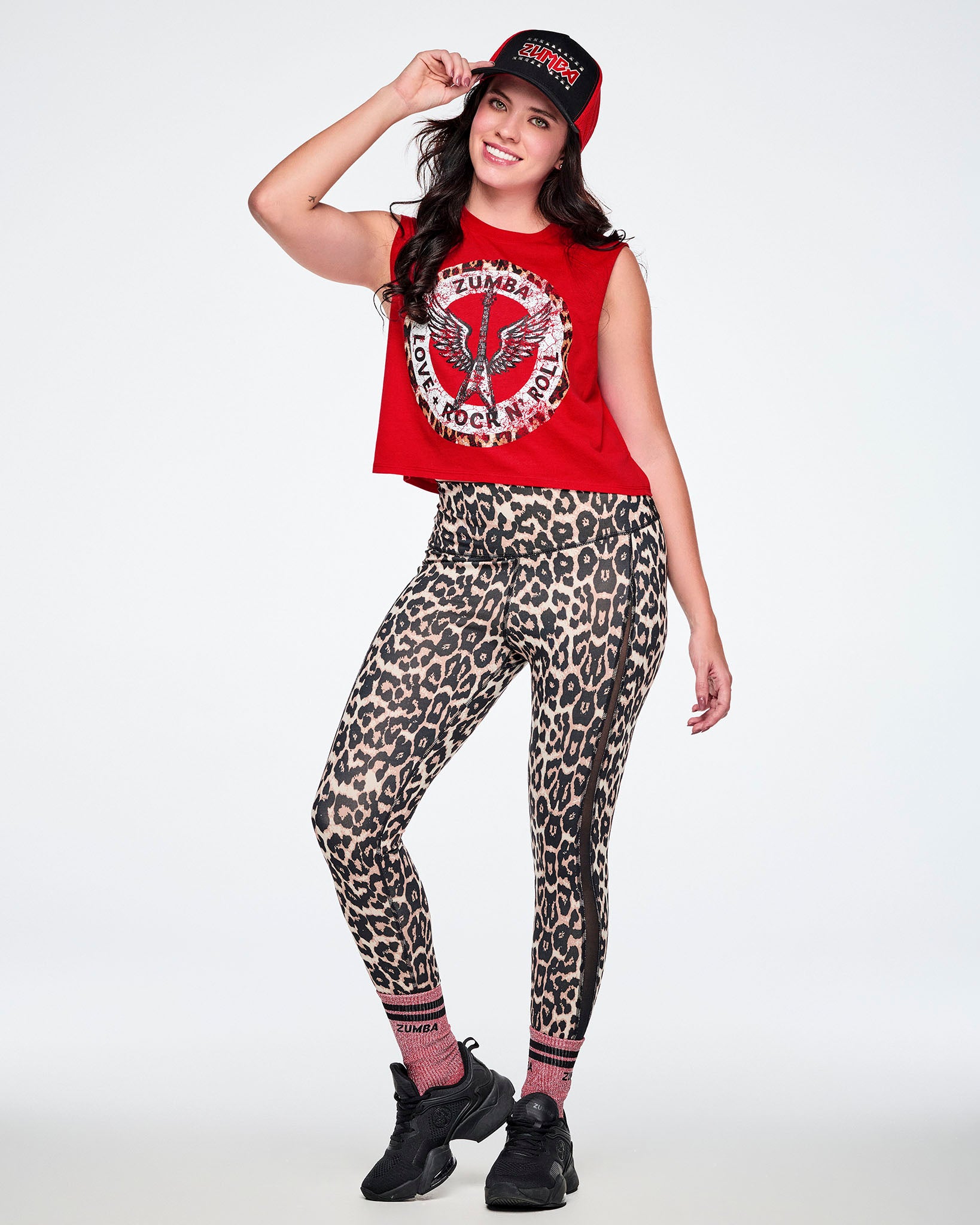 A female model modeling a red and black Zumba hat, a red Zumba rock graphic crop top, leopard-print leggings paired with black shoes.
